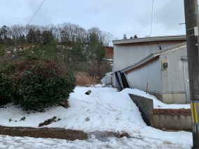雪の残る庭と、傾いた塀のある住宅